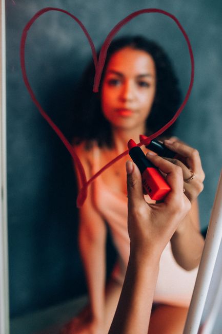 A woman uses red lipstick to draw a heart shape on a mirror, seen in reflection.