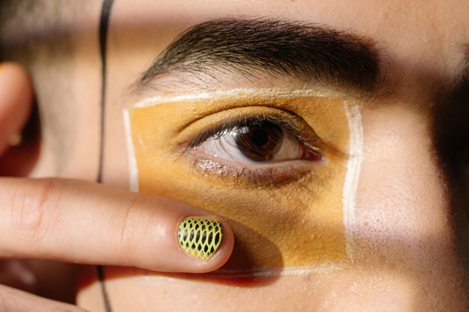 Artistic close-up of an eye with creative makeup and detailed nail art in bright lighting.