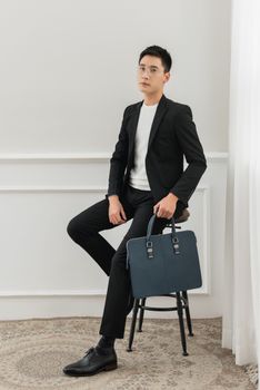 Portrait of a professional Asian man sitting on a stool, elegantly dressed in a suit holding a stylish briefcase.