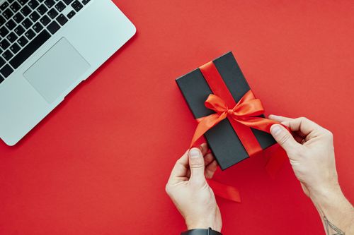 Free Close-up of hands tying a red ribbon on a gift box beside a laptop on a red background, perfect for holiday presents. Stock Photo