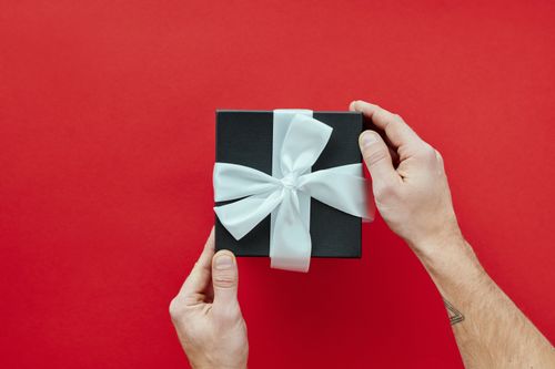 Free Elegant black gift box with a white ribbon held by hands against a vibrant red background. Stock Photo