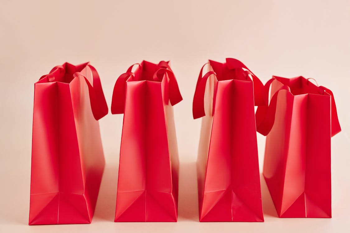 Line of red gift bags with ribbon handles on a light pink background.