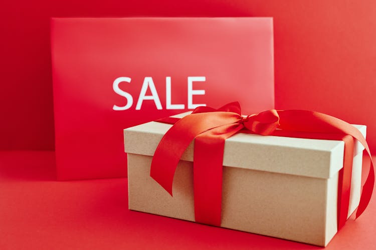 Cardboard Box With Red Ribbon Beside A Sale Sign