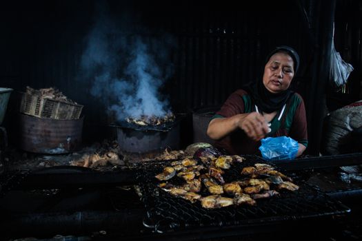 An elderly woman grills chicken over charcoal in a traditional setting, with smoke rising.