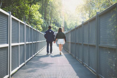 Free A couple holding hands walking along a forest bridge surrounded by lush greenery. Stock Photo
