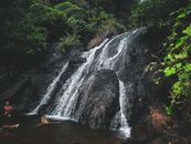 Unrecognizable travelers chilling in warm pond formed by waterfall flowing through rough stony cliff in lush forest