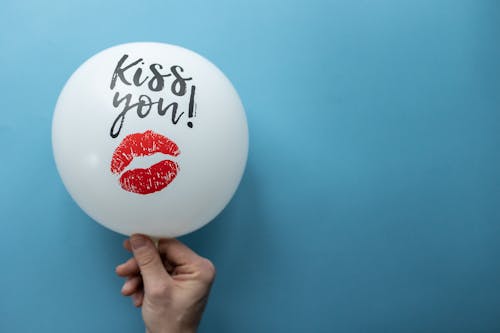 Unrecognizable person demonstrating white balloon with Kiss You inscription and kiss mark on blue background in studio during holiday celebration