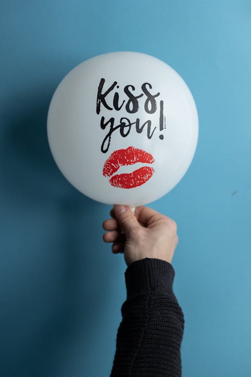 Unrecognizable male in black sweater showing white balloon with Kiss You lettering and lipstick kiss mark on blue background during holiday celebration