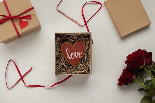 Top view of heart shaped cookie with Love word in box with decorative paper filling [laced on gray background near gift boxes with red ribbons and bouquet of roses