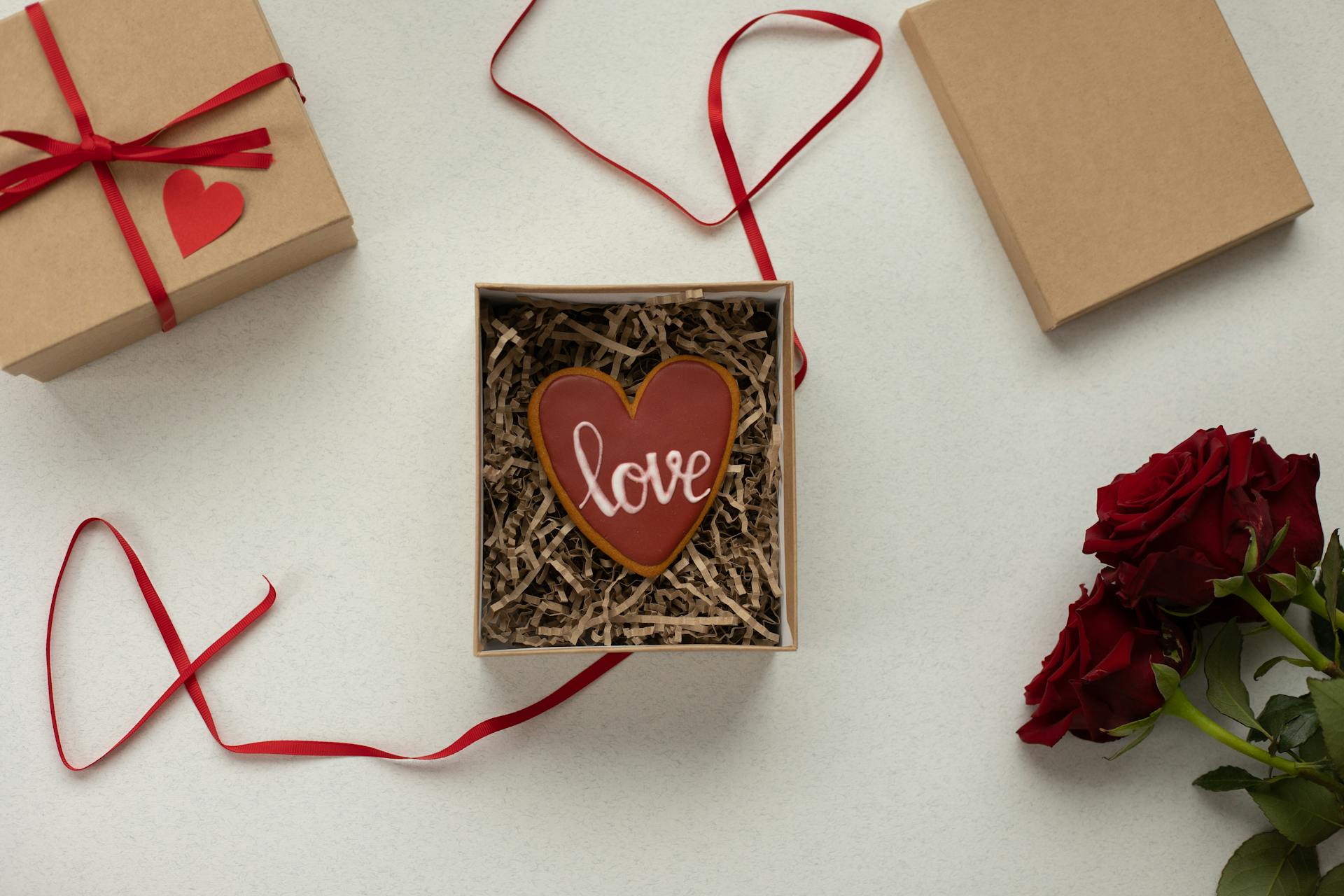 Top view of heart shaped cookie with Love word in box with decorative paper filling [laced on gray background near gift boxes with red ribbons and bouquet of roses