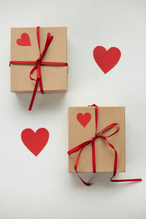 Top view of cardboard boxes tied with ribbons placed on white background with paper red hearts during saint valentines day