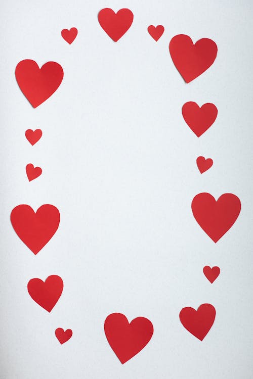 Top view paper cutout of different red hearts arranged in shape of oval on white background during saint valentine day