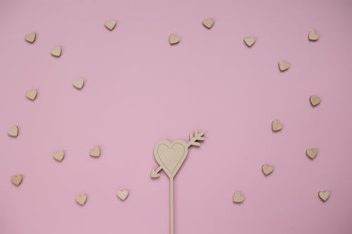 From above of composition of wooden heart with arrow surrounded by small wooden hearts against pink background