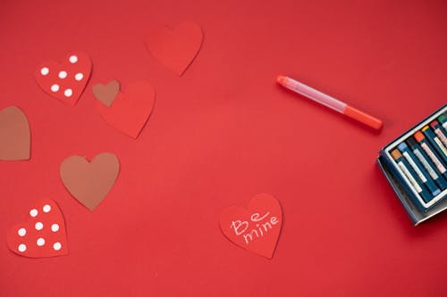 Top view of Be Mine inscription written on paper heart and placed on red background with felt pens during valentines day
