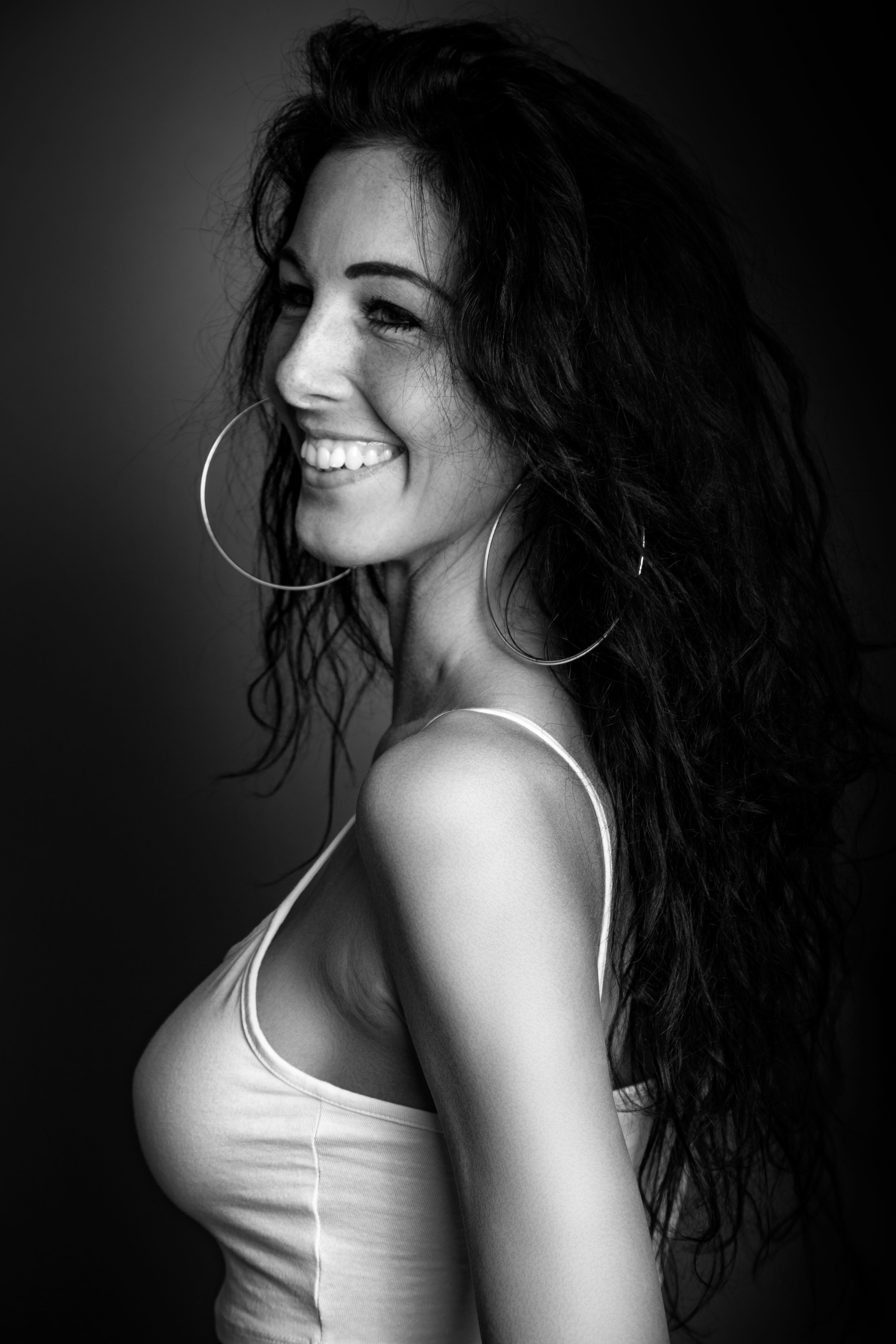 A captivating monochrome portrait of a woman smiling, wearing hoop earrings and a spaghetti strap top.