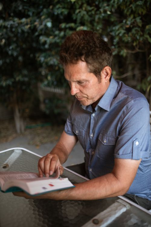 Free Adult man reading a book outdoors at a table surrounded by greenery. Stock Photo