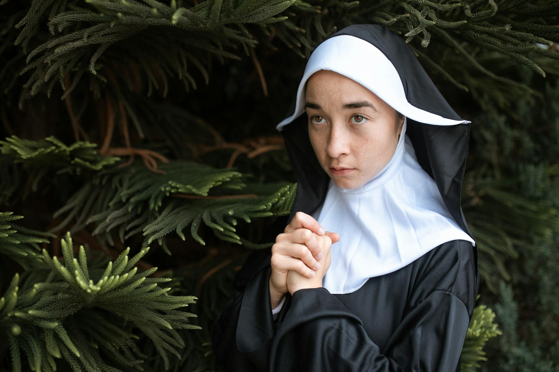 A young caucasian nun in prayer poses outdoors surrounded by green foliage, conveying peace and faith.