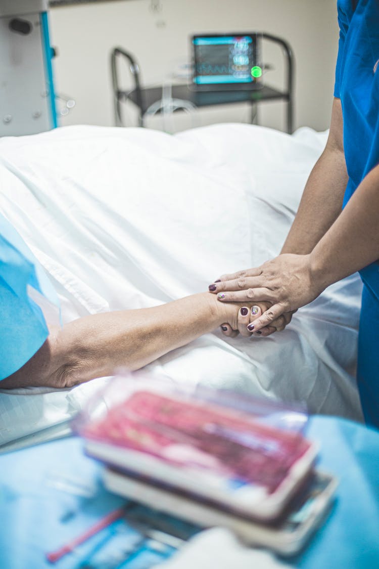 Person In Blue Scrubs Holding The Hand Of A Patient  Lying On Bed