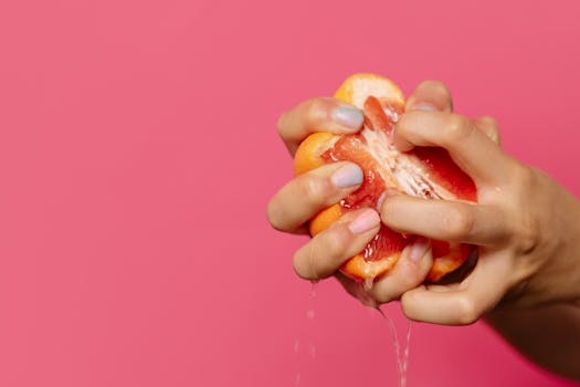 Close-up of hands squeezing juicy grapefruit against a bright pink backdrop, emphasizing freshness.