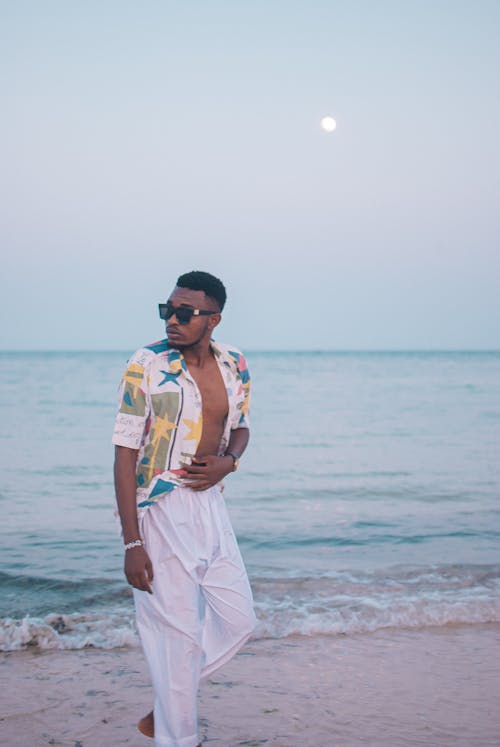 Self assured young barefooted African American male tourist in stylish summer outfit and sunglasses walking on sandy beach near wavy ocean at sunset