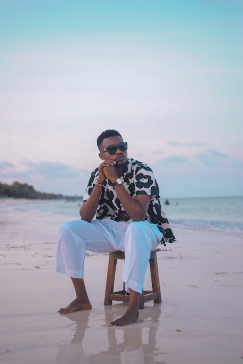 Pensive young black man relaxing on stool on sandy seacoast against sunset sky