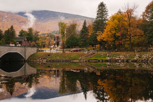Free Heavy clouds over mountain ridge and vibrant green and orange trees reflecting from water surface of calm lake in town in autumn Stock Photo