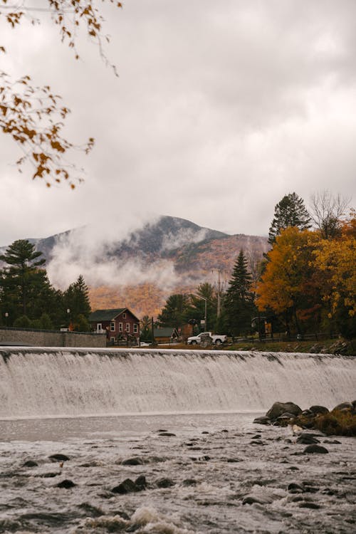 Dense clouds and haze over tall mountain and green and yellow trees next to bridge over river dawn in daytime