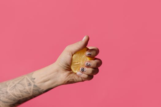 Tattooed hand squeezing a lemon against a vibrant pink background, emphasizing contrast and texture.