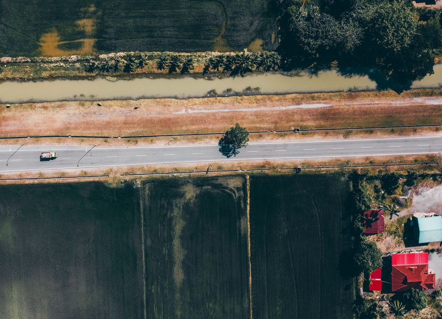 Aerial view of straight roadway with driving vehicle between green plantations near house roofs in daylight