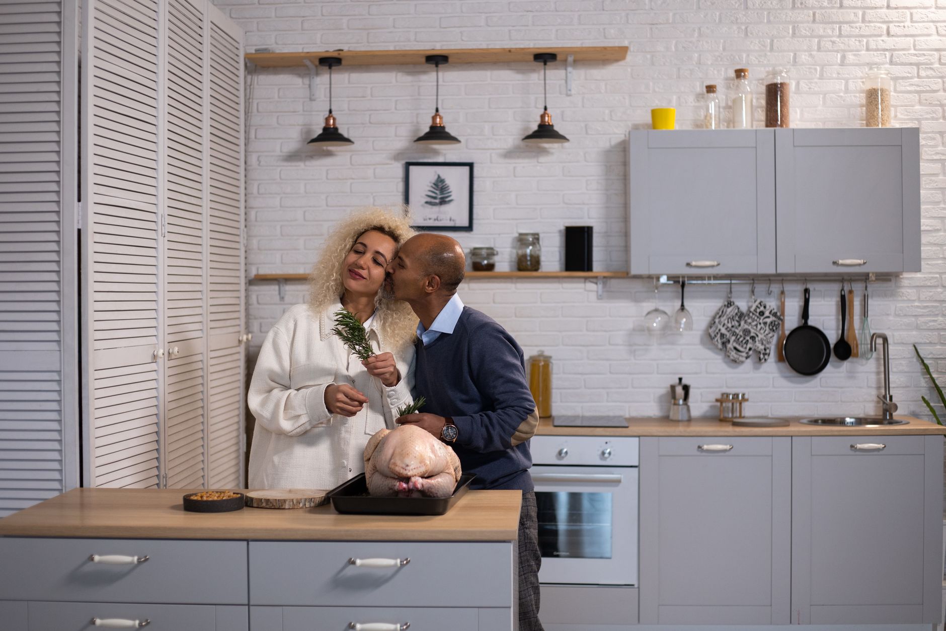 Romantic African American husband kissing cheek of black wife with rosemary while standing at counter with raw turkey in kitchen
