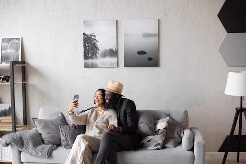 Smiling Asian woman and African American man sitting on comfortable couch and having video call on cellphone in modern living room