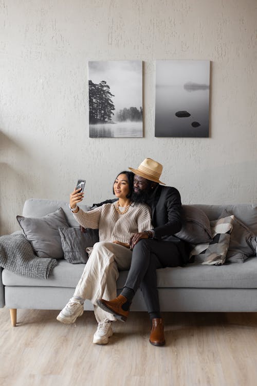 Full body of content Asian woman and African American man having video call while sitting on couch with crossed legs in room