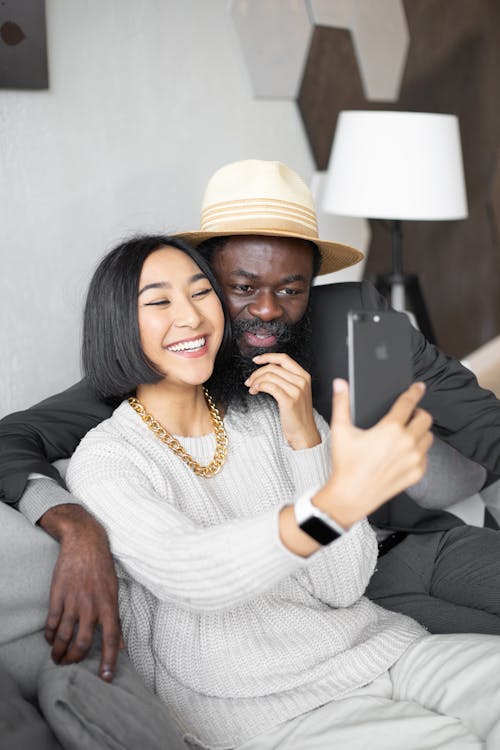 Happy Asian woman and African American man having video chat on modern cellphone while sitting on couch in modern room