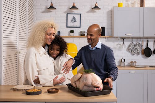 Positive African American parents with daughter hugging while standing at table with raw turkey while cooking together in modern kitchen