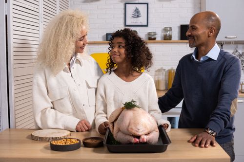Free Content African American parents with daughter looking at each other while standing at counter with uncooked turkey and plates with ingredients Stock Photo