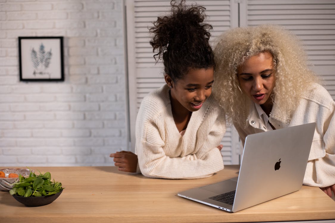 Positive African American mother and daughter looking at screen of modern netbook while standing at wooden table with fresh food