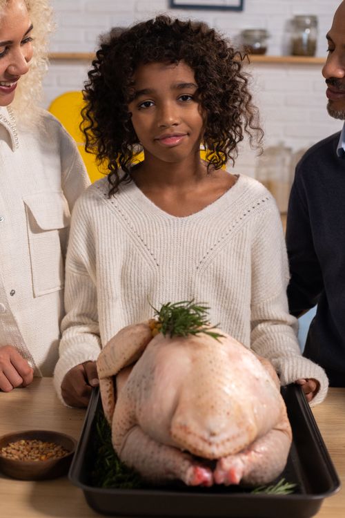 Free African American daughter with tray of raw turkey looking at camera while standing at counter with anonymous parents in kitchen Stock Photo