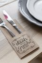 From above of metal shiny fork and knife placed in paper cover with inscription for Thanksgiving near plates on wooden table