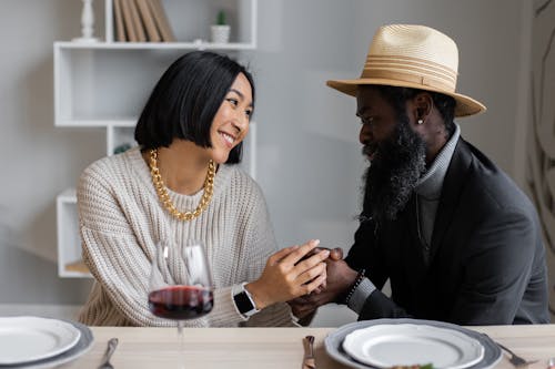 Loving multiracial couple looking at each other holding hands while sitting at table with plates and wine and having dinner