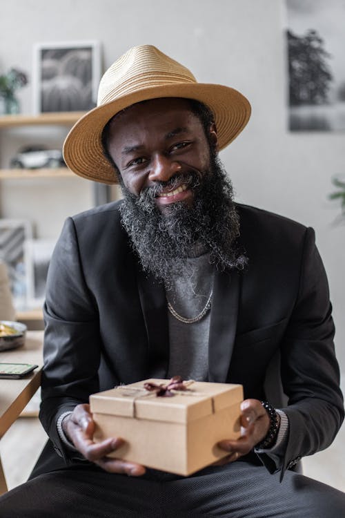 Cheerful adult African American male with beard in jacket and hat with carton gift box looking at camera