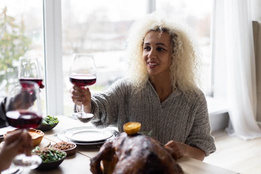 Delighted ethnic female clinking glasses of wine with guests while sitting at table with roasted turkey and having dinner on Thanksgiving Day
