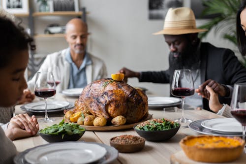 Calm multiracial friends holding hands with closed eyes sitting at table served with roasted turkey and other dishes and praying before having Thanksgiving dinner