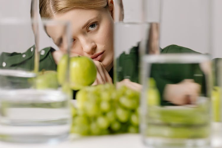 Woman In Green Long Sleeves Near Green Fruits And Glasses Of Water While Seriously Looking At The Camera