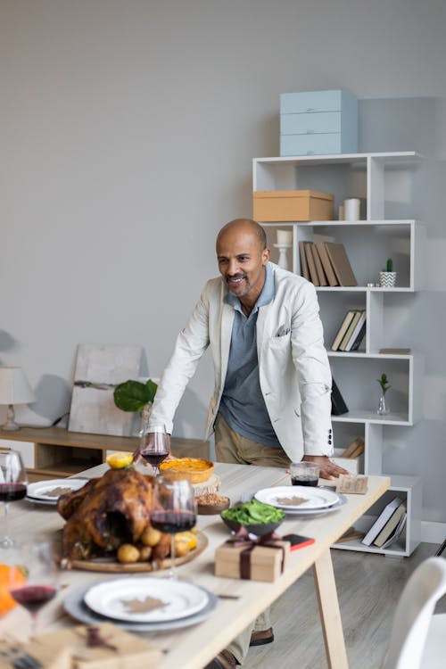 Positive bearded bald adult Indian man in casual clothes leaning on table served with wine and roasted turkey for celebrating Thanksgiving Day