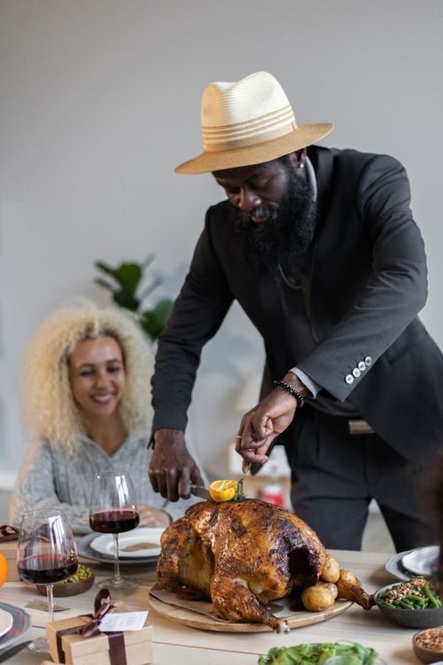 Free Positive bearded African American male in suit and hat cutting roasted turkey with knife and fork while having Thanksgiving dinner with guests Stock Photo