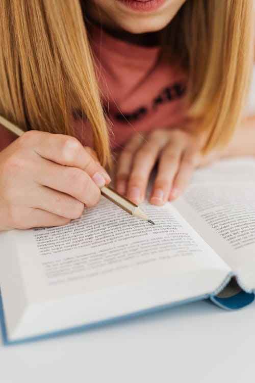 Close Up Photo of a Person Holding Pencil While Reading a Book