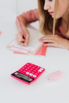 Focus on a woman writing in a notepad with a pink calculator and eraser nearby.
