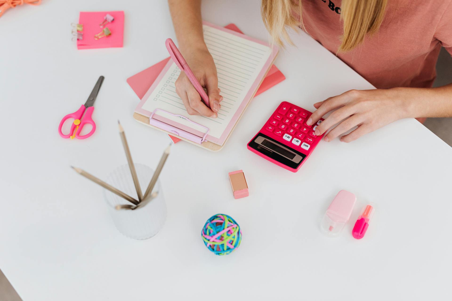 A woman using pink stationery on a desk, including a calculator and notepad. Bright and organized workspace.