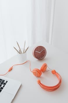 A minimalist desk setup featuring a clock, headphones, and a laptop in orange accents.