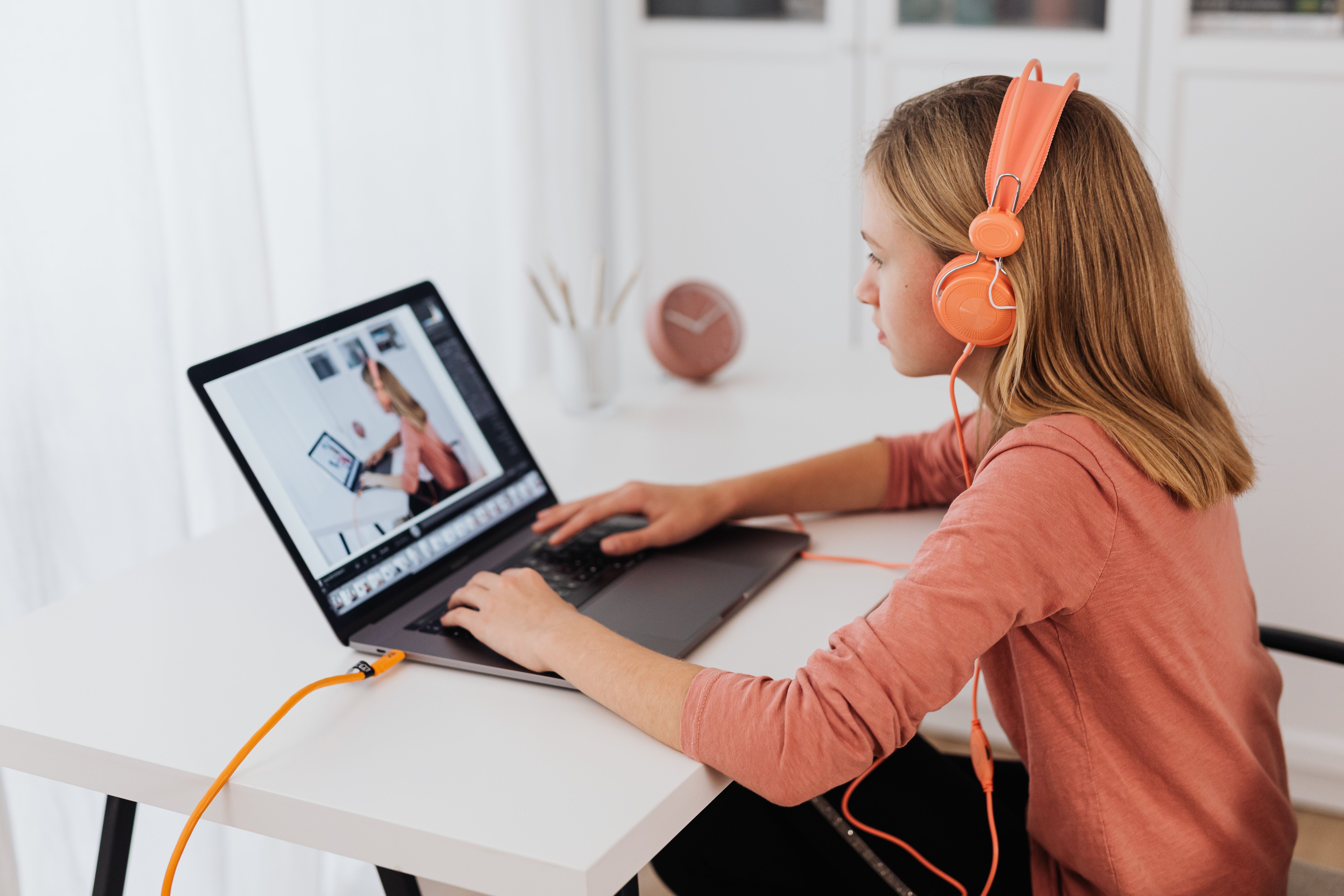 Teen girl with orange headphones engaged with laptop on a desk in a home setting.